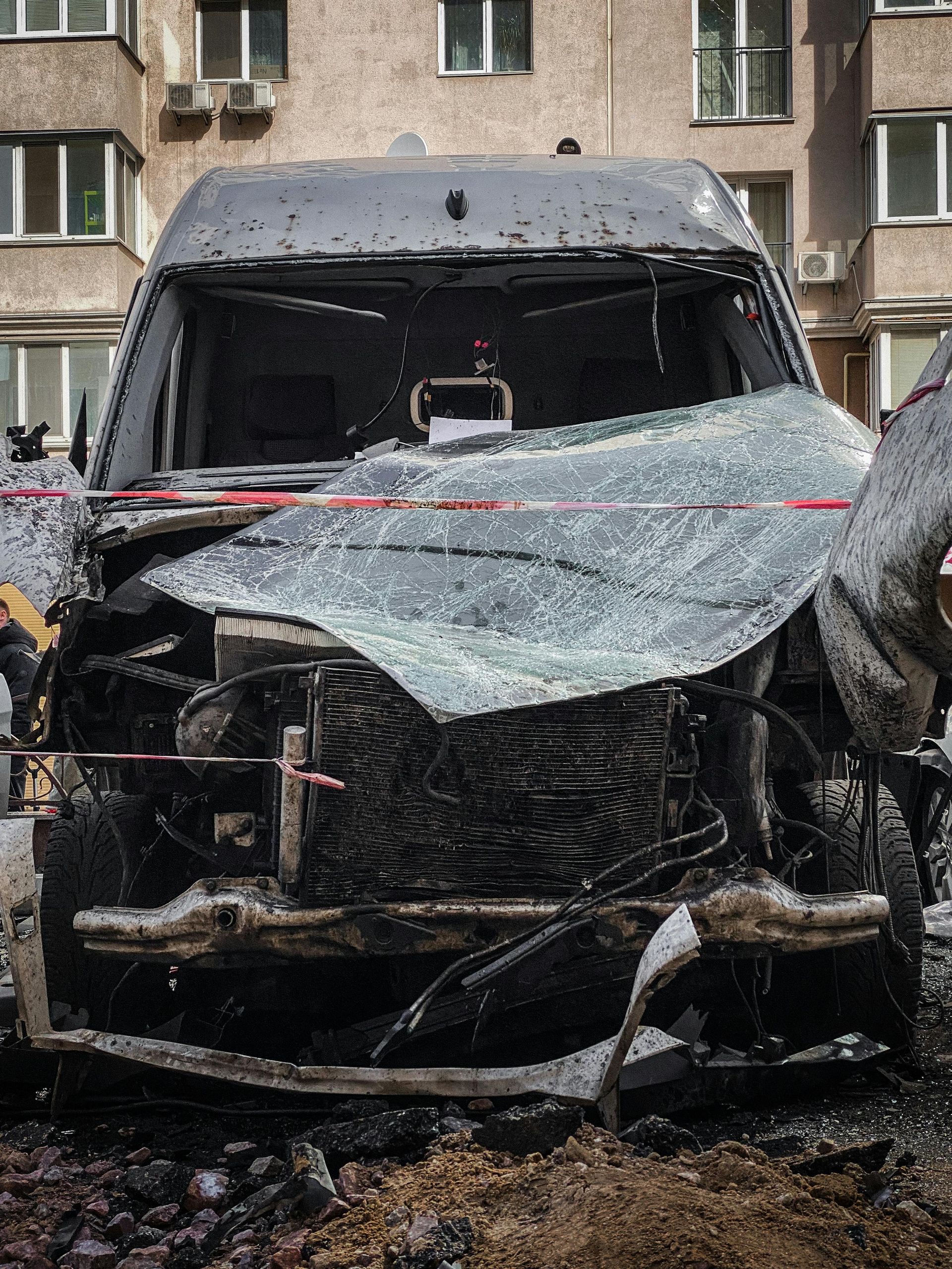 A severely fire-damaged, charred vehicle sits parked in front of an urban apartment building.