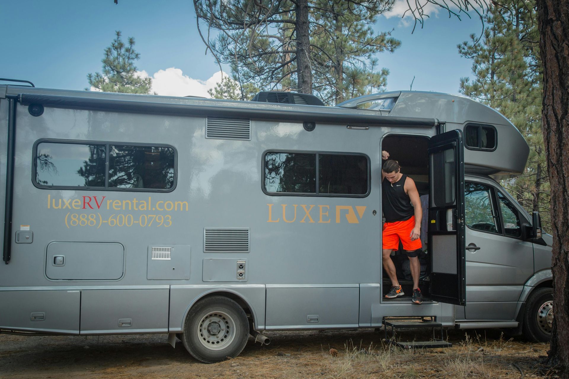 A man in a black tank top and orange shorts steps out of a grey Luxe RV parked in a forest setting.