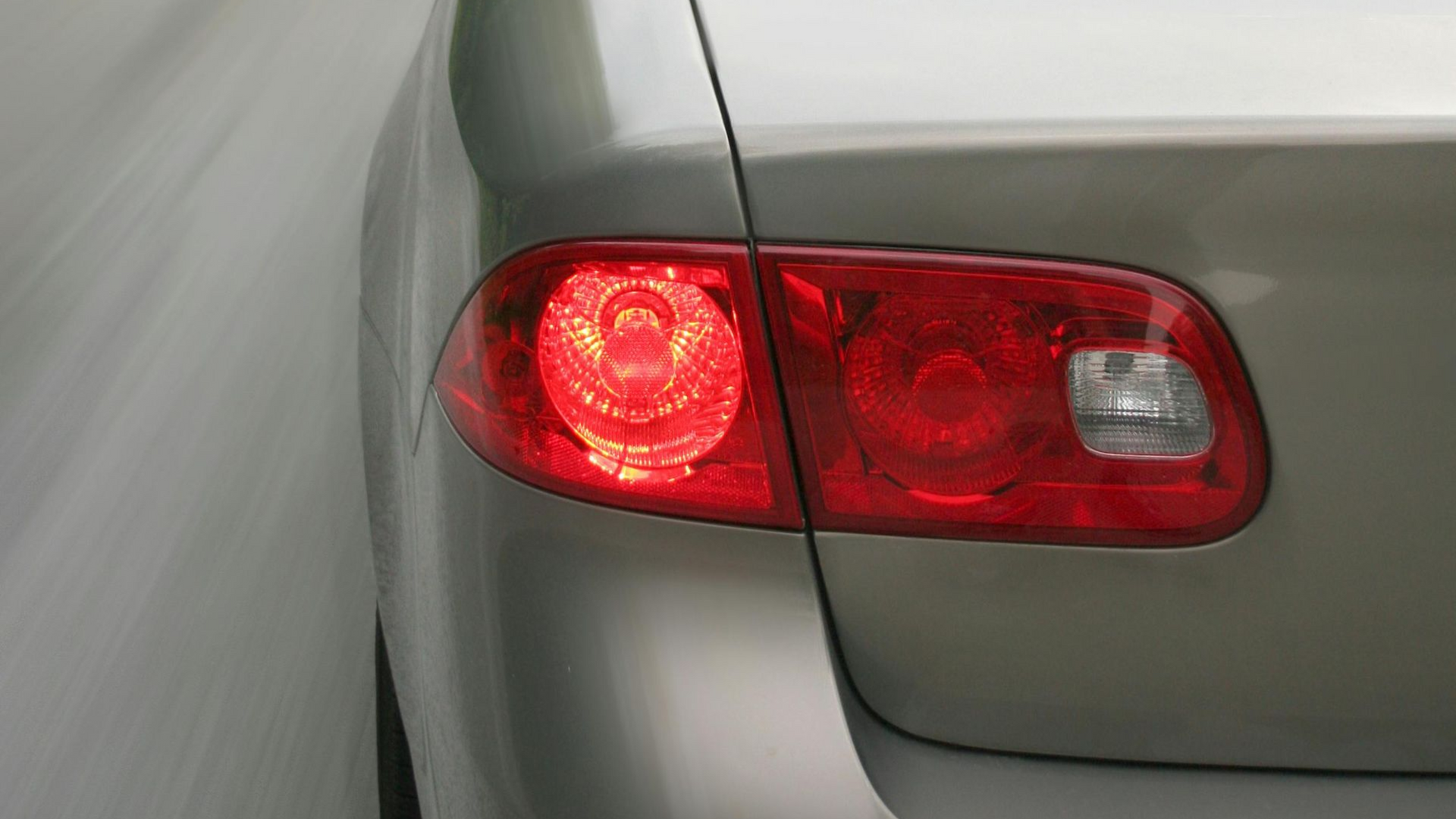 Rear of a silver car with a glowing red left taillight on a blurred road.