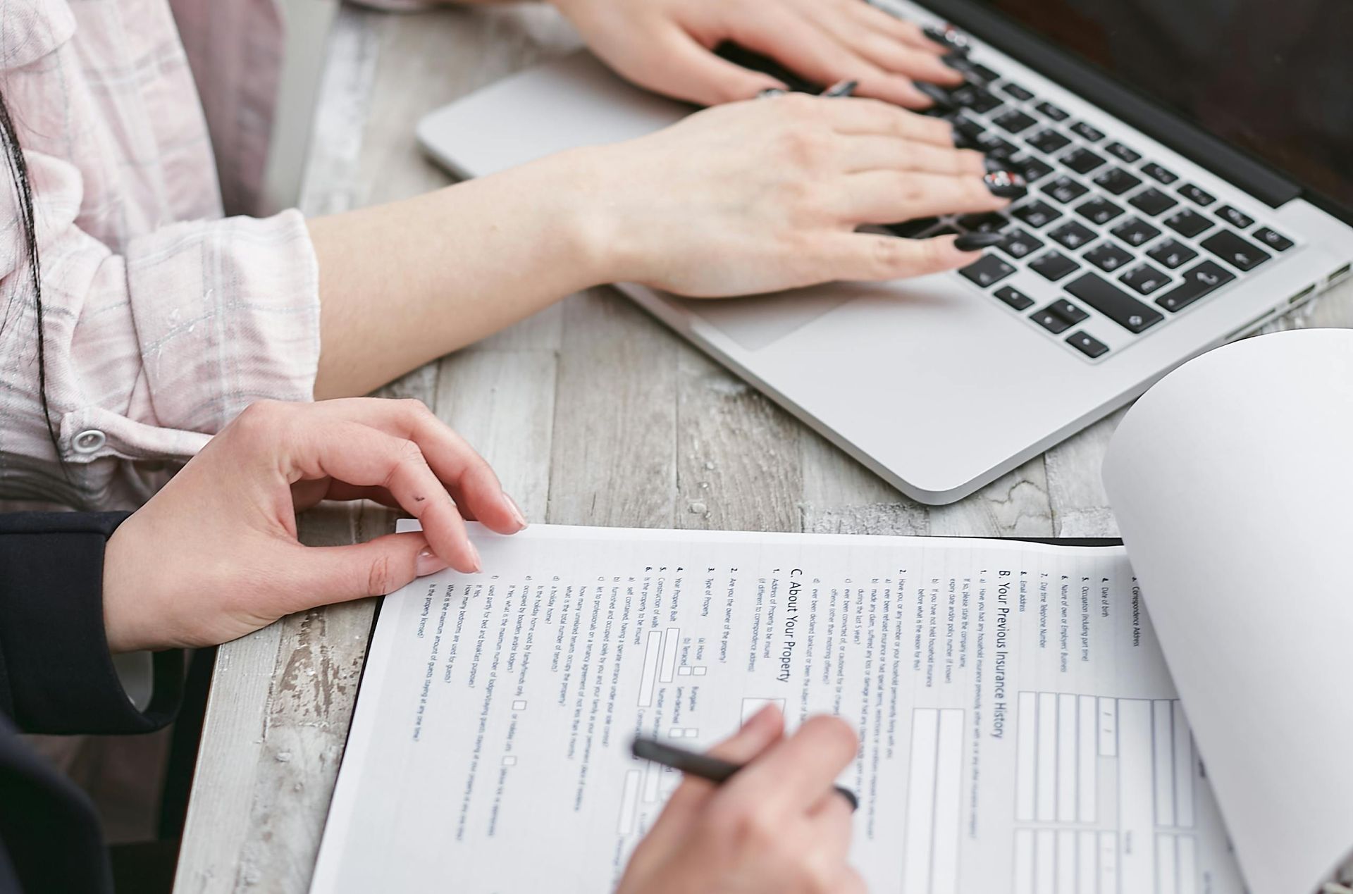 Two people working together at a desk, one typing on a laptop and the other reviewing a printed document with a pen.