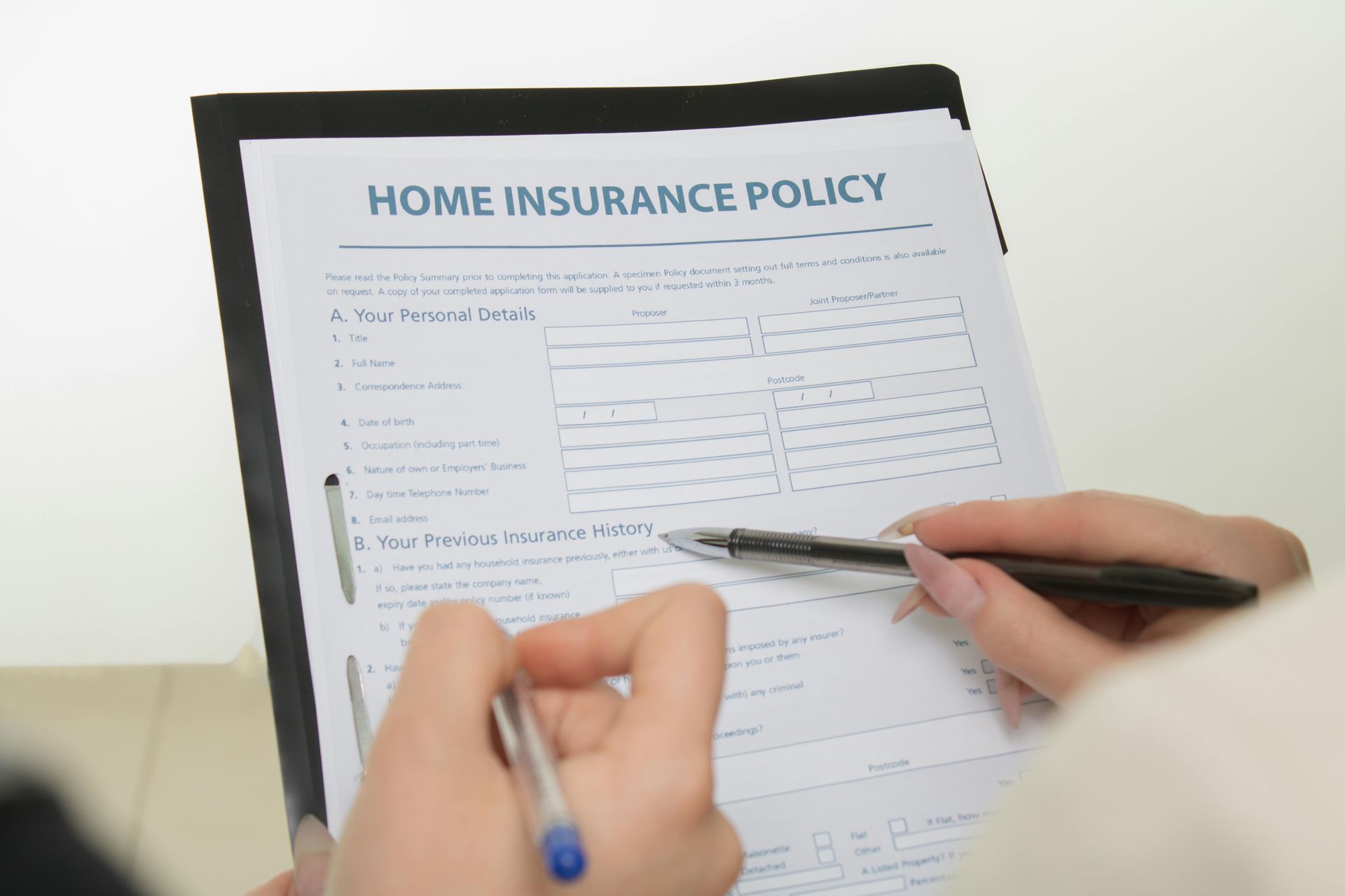 Two hands use pens to fill out a home insurance policy form on a clipboard.