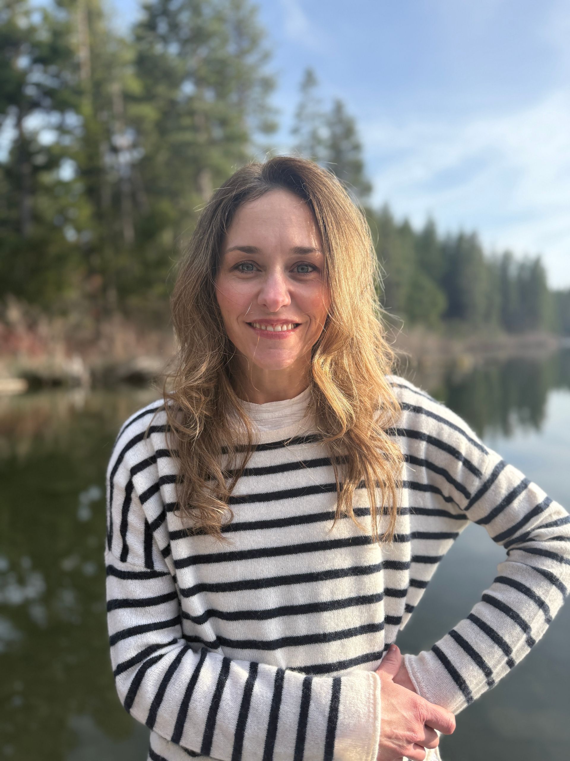 Woman stands and smiles in front of trees wearing blue and white striped sweater