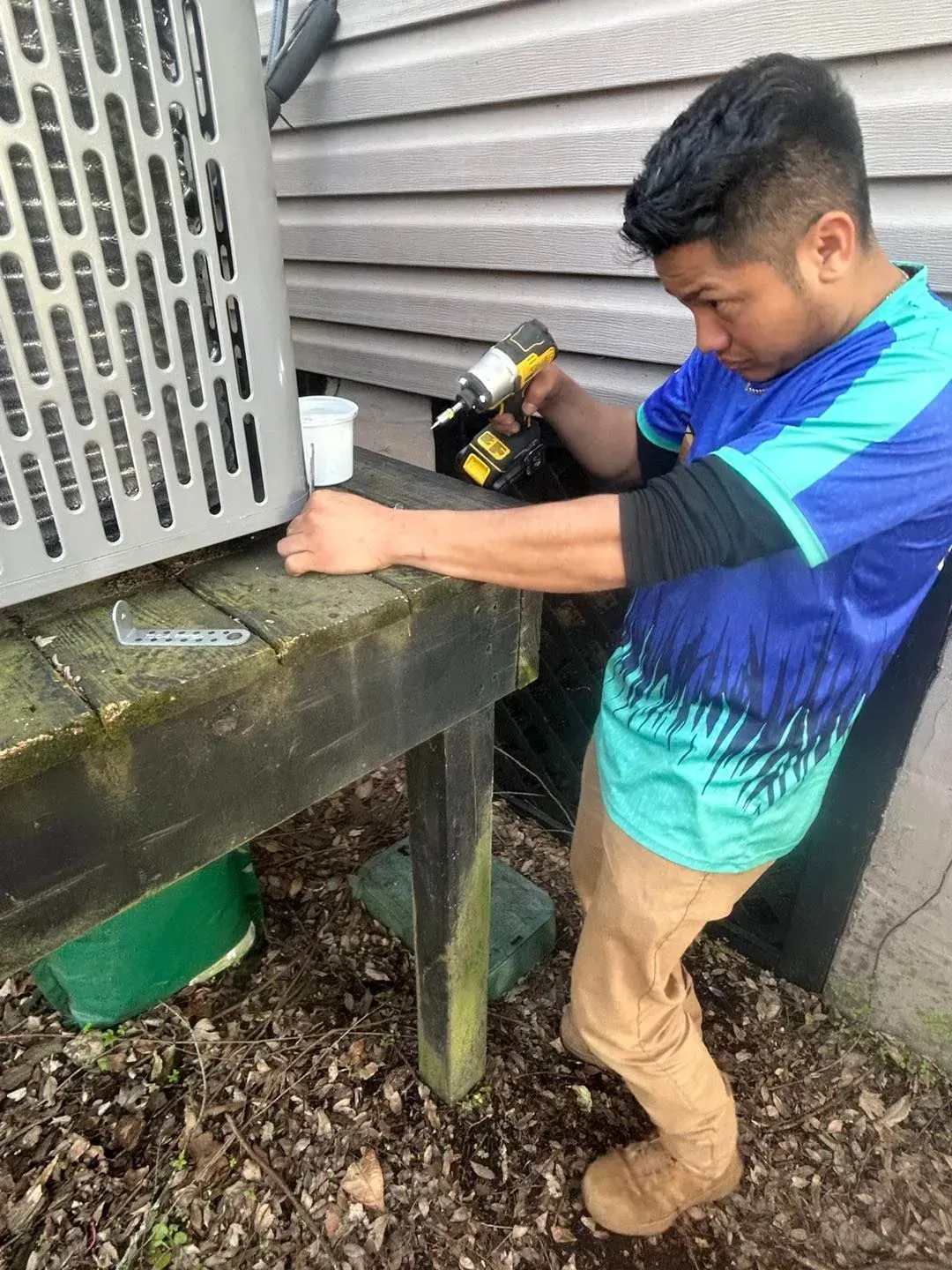 Man working on an outdoor air-conditioning unit with a drill beside a house