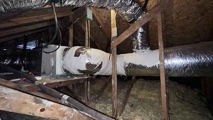 Damaged white ductwork in an attic with exposed wooden beams and insulation.