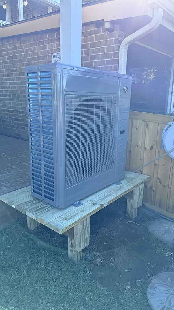 Outdoor HVAC unit on a wooden platform beside a brick wall, with a vented side panel and visible frost or dust.