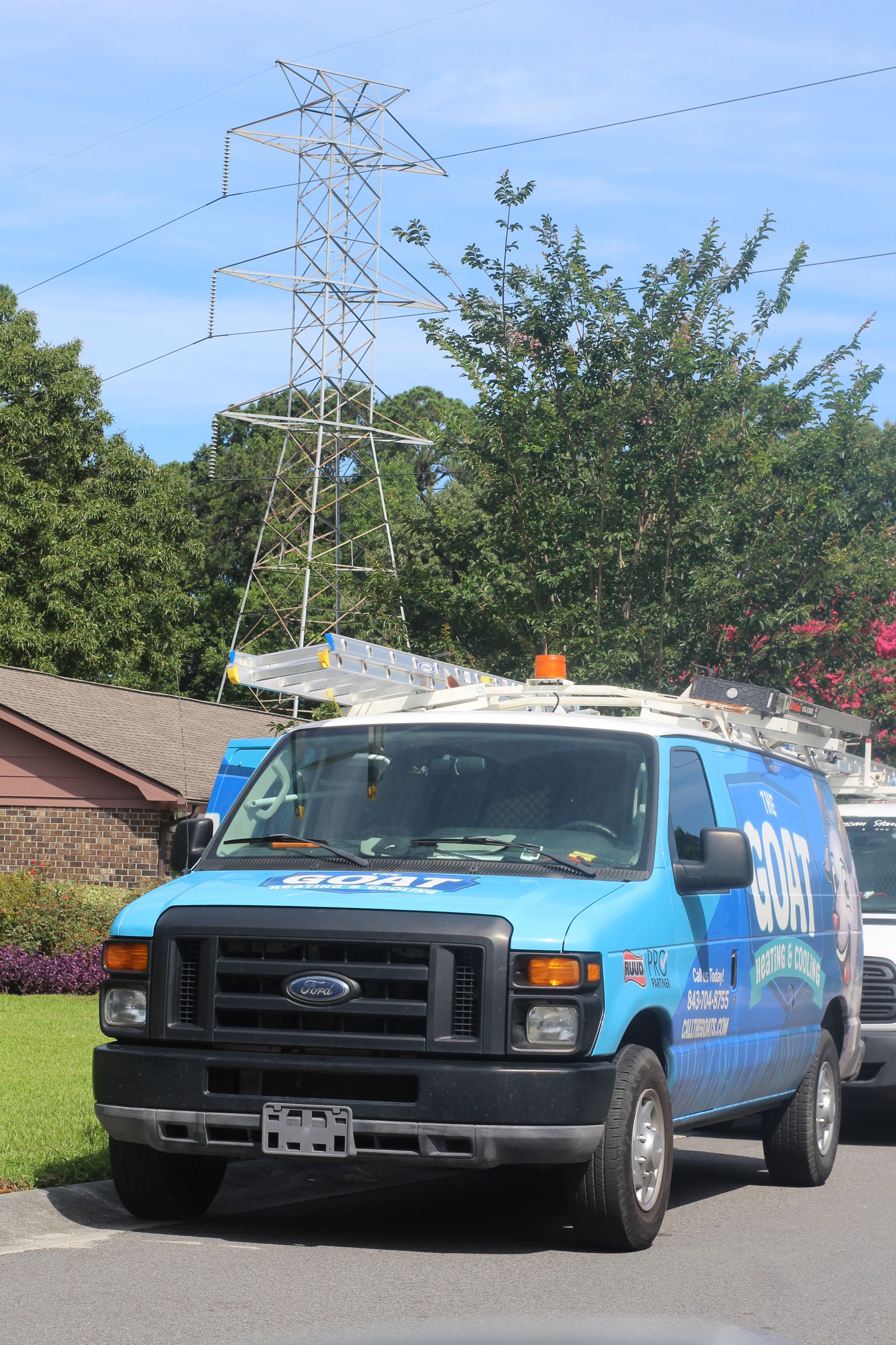 Blue utility van parked on a road beside greenery and power lines under a bright sky