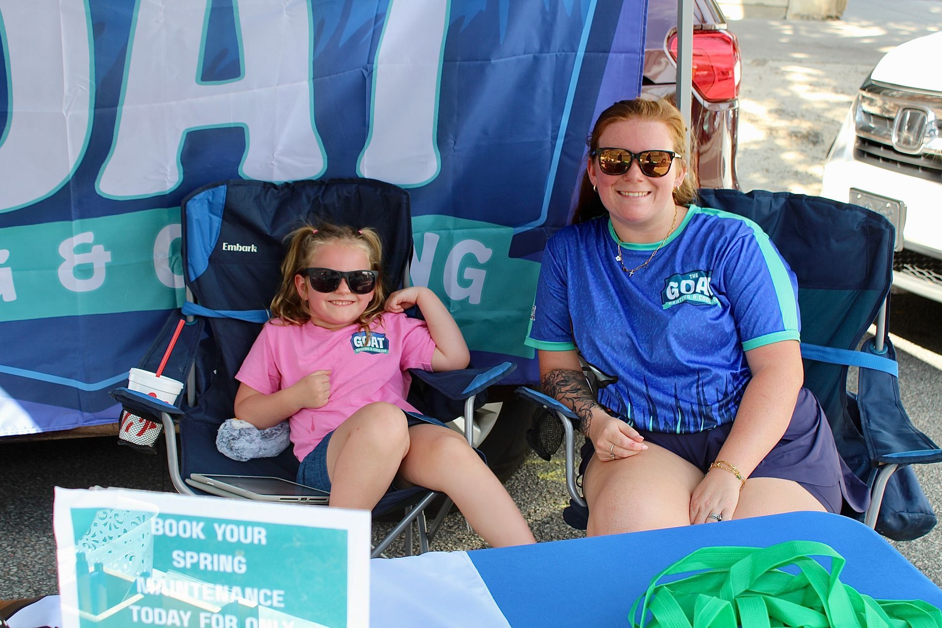 Two people sitting under a blue tent, smiling at an event booth with a sign in front.