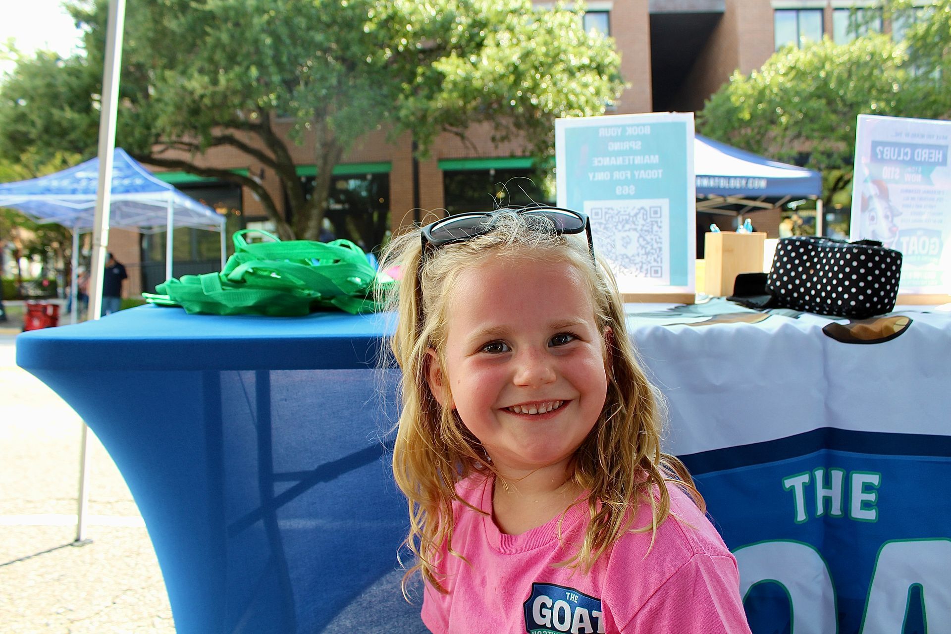 Smiling child in pink shirt at an outdoor booth with blue tablecloth and tents in the background