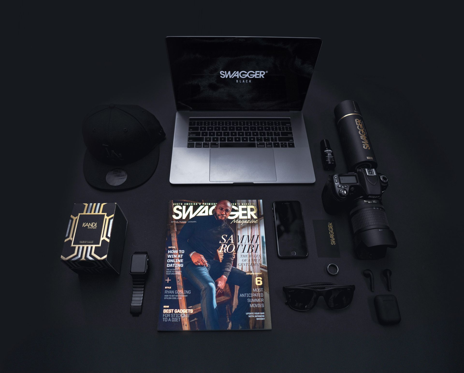 Flat lay of a laptop, magazine, cap, camera gear, and gadgets on a dark surface