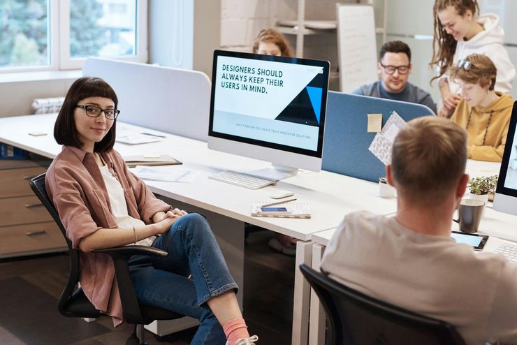 People in a meeting around a large table, with a woman in glasses and a presentation screen in a bright office.