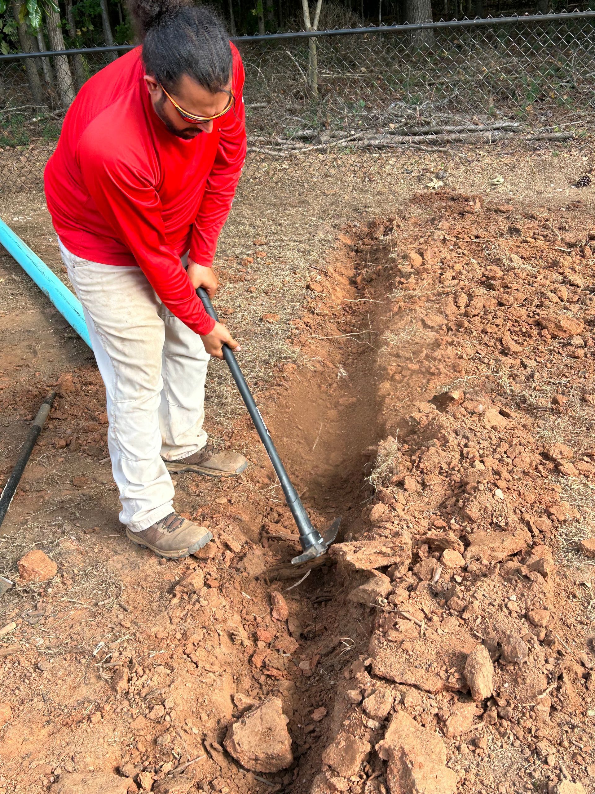 A person in a red shirt digs a trench in brown soil with a pickaxe outdoors.