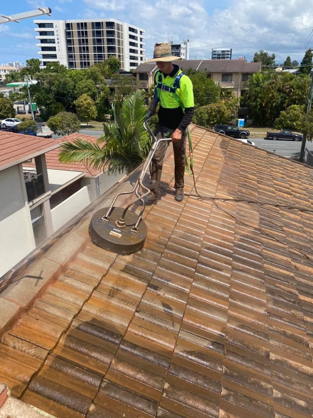 A man cleaning a red tile roof with a pressure washer on a sunny day, city in the background — SEQ Roofing & Painting in Nerang, QLD