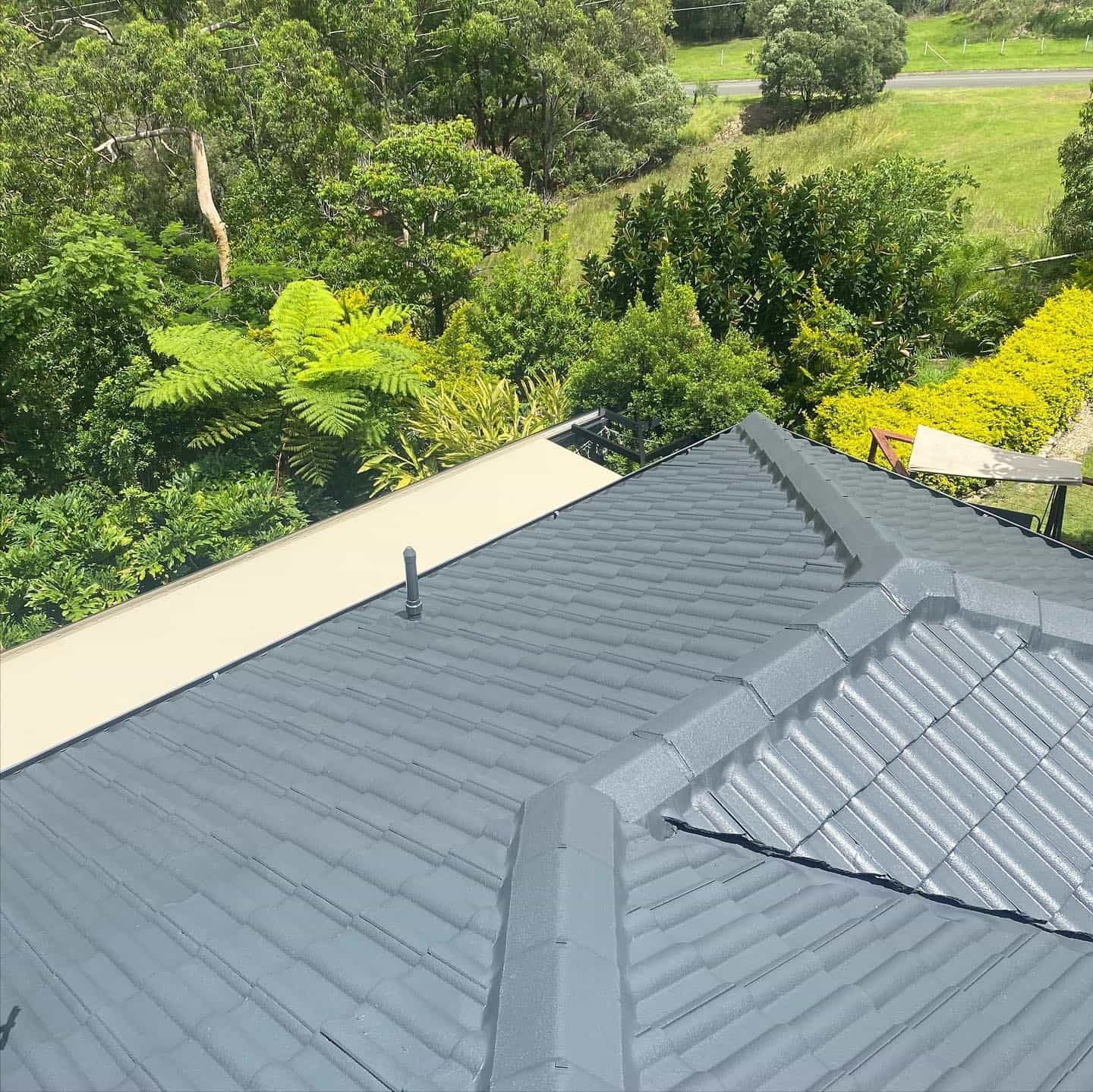 Grey Metal Roof With Angled Ridges, Set Against Green Trees and a Sunny Sky — SEQ Roofing & Painting in Mudgeeraba, QLD