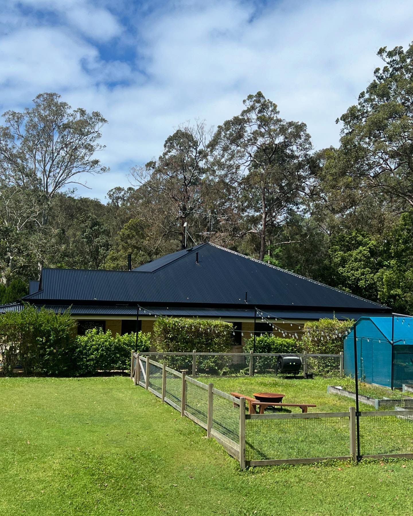 House With Dark Blue Roof, Fenced Yard, and Surrounding Trees Under a Cloudy Sky — SEQ Roofing & Painting in Carrara, QLD