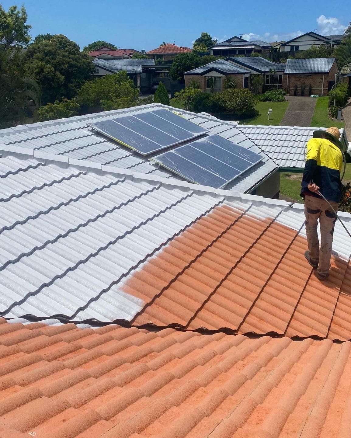 A worker sprays a terracotta roof white near solar panels. Homes are in the background. — SEQ Roofing & Painting in Nerang, QLD