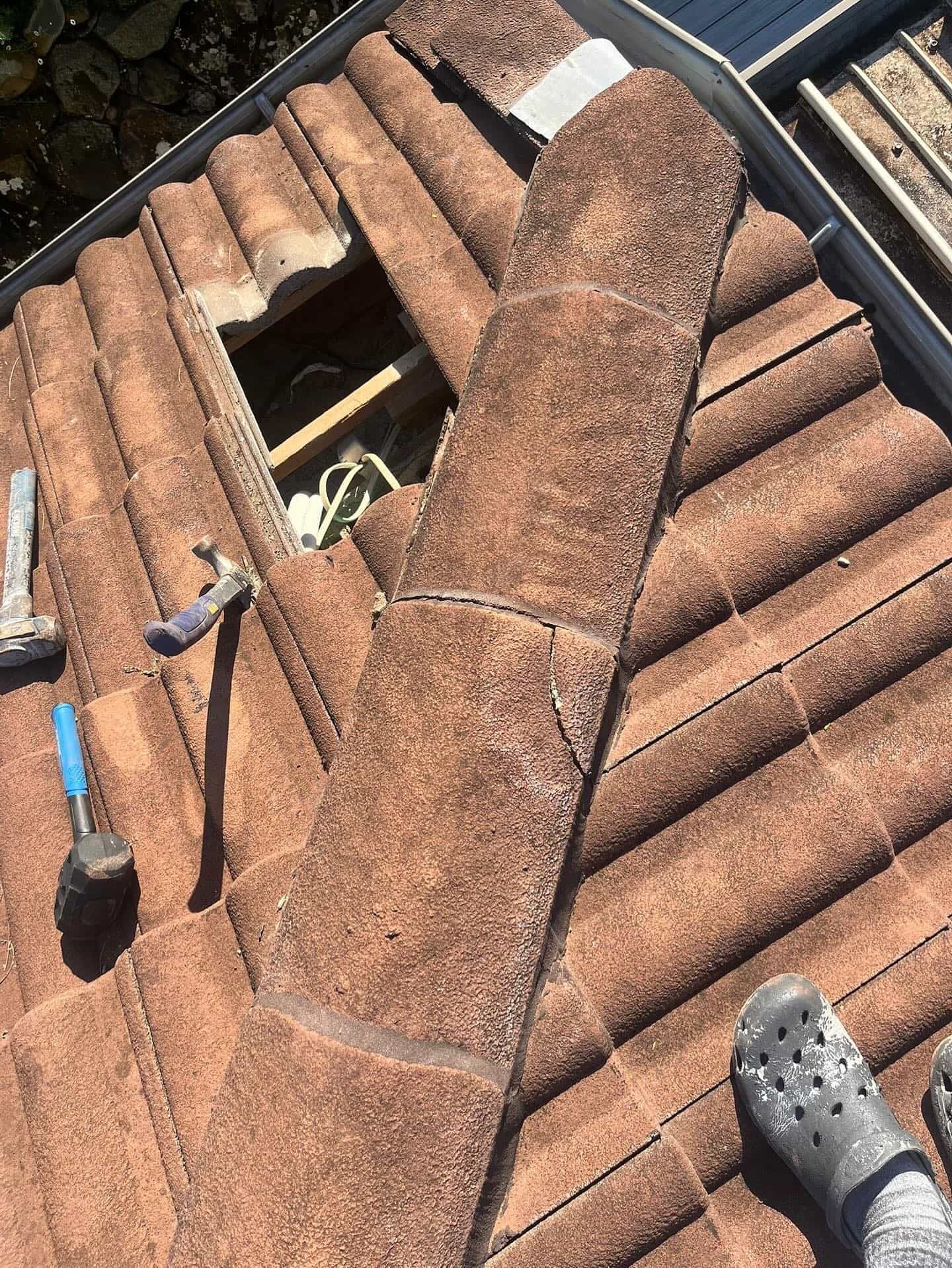 Rooftop with missing tiles exposing interior. Brown tiles, tools, and a foot wearing a Croc —SEQ Roofing & Painting in Nerang, QLD
