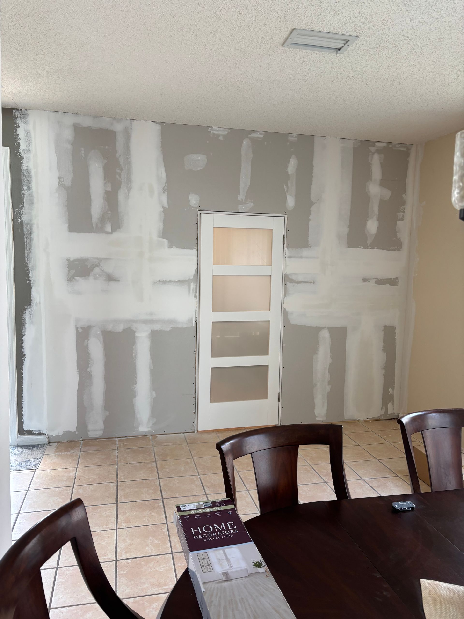 Living room with a gray drywall wall under renovation and a dark dining table in the foreground.