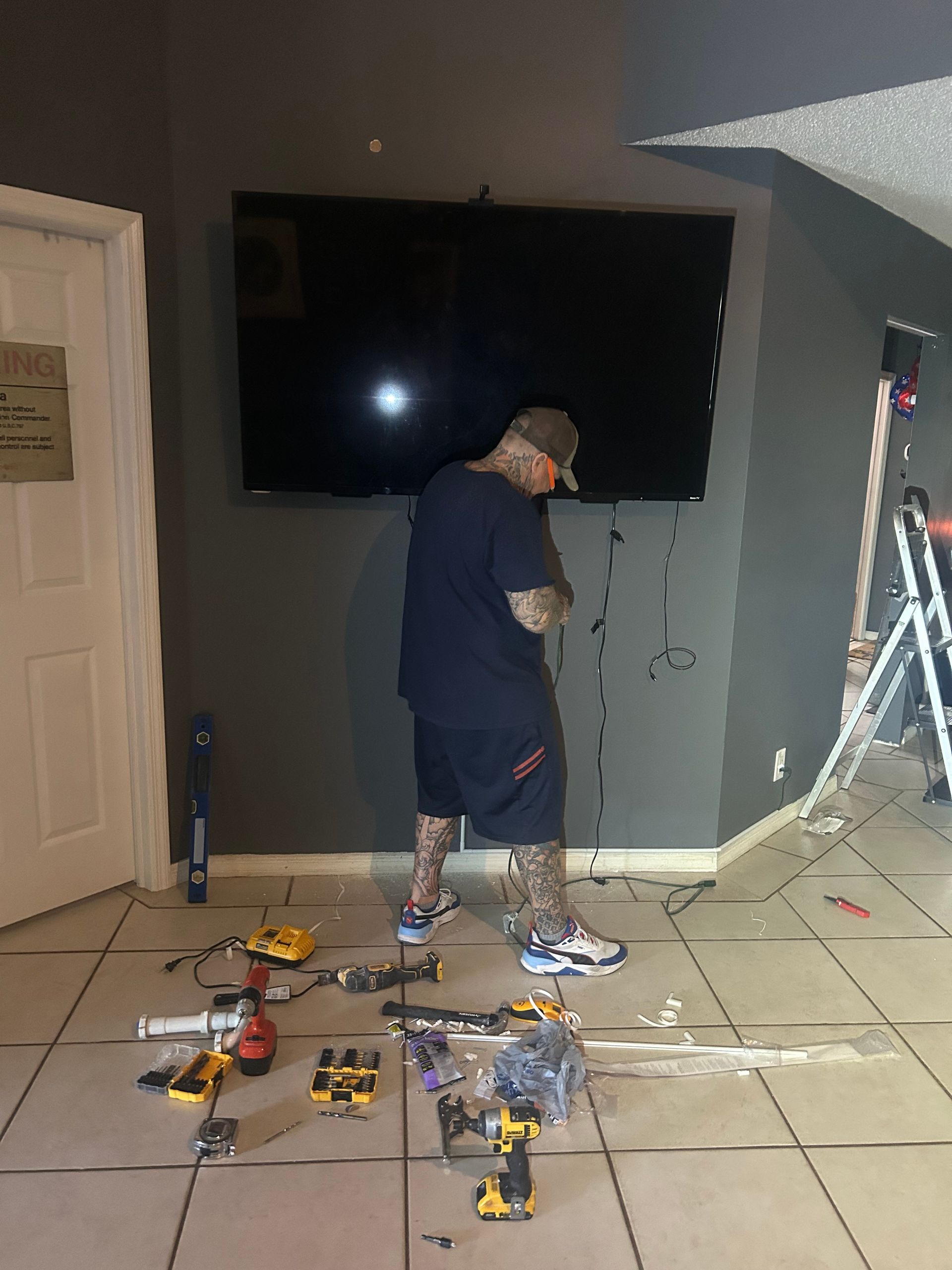 Person working amid tools and debris while mounting a large TV on a gray wall in a room under renovation.