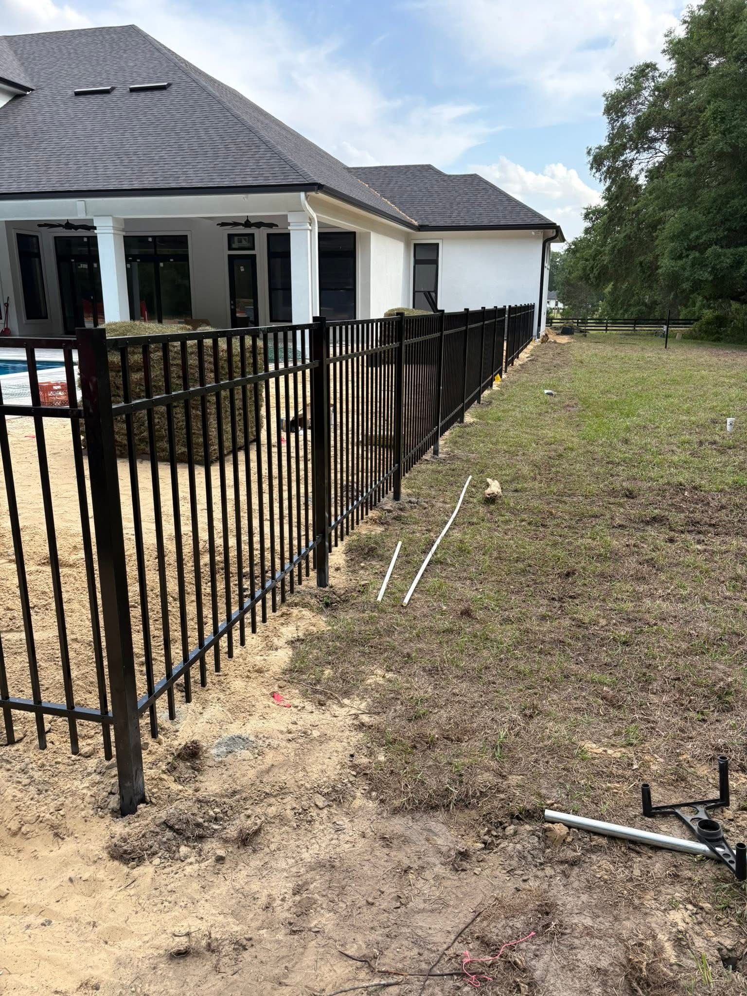 Black metal fence beside a white house on a dirt lot with scattered construction pipes and trees nearby
