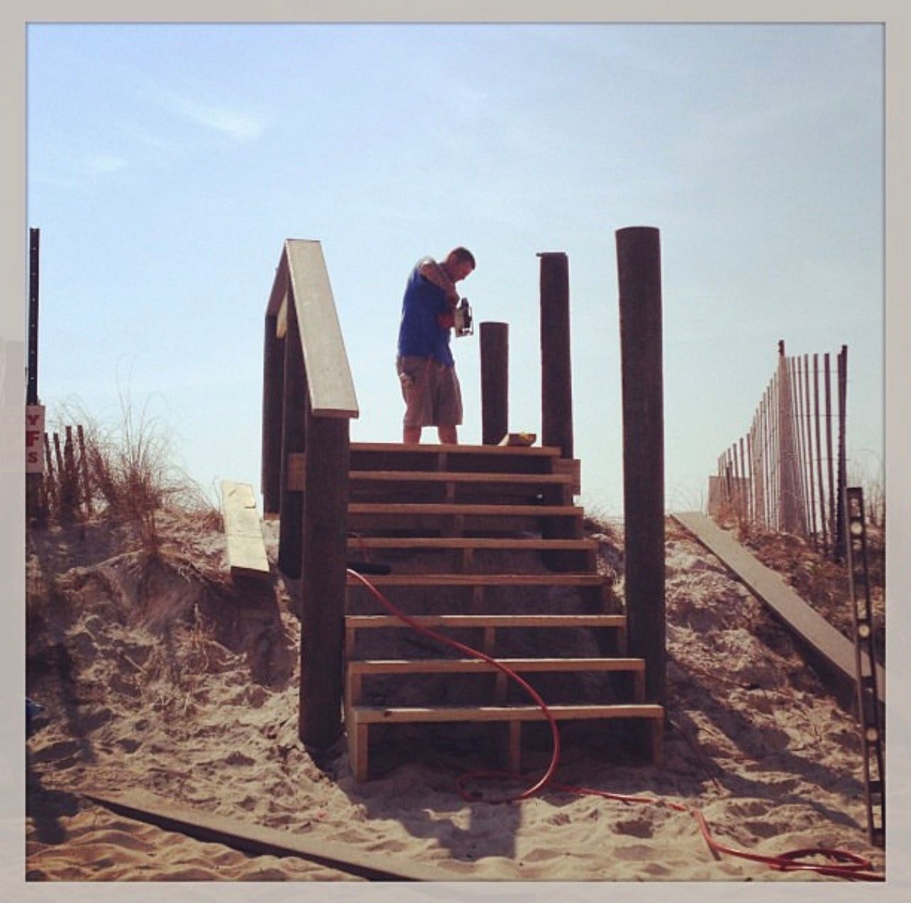Person standing on wooden beach stairs, working near tall posts and a fence in a sandy dune area