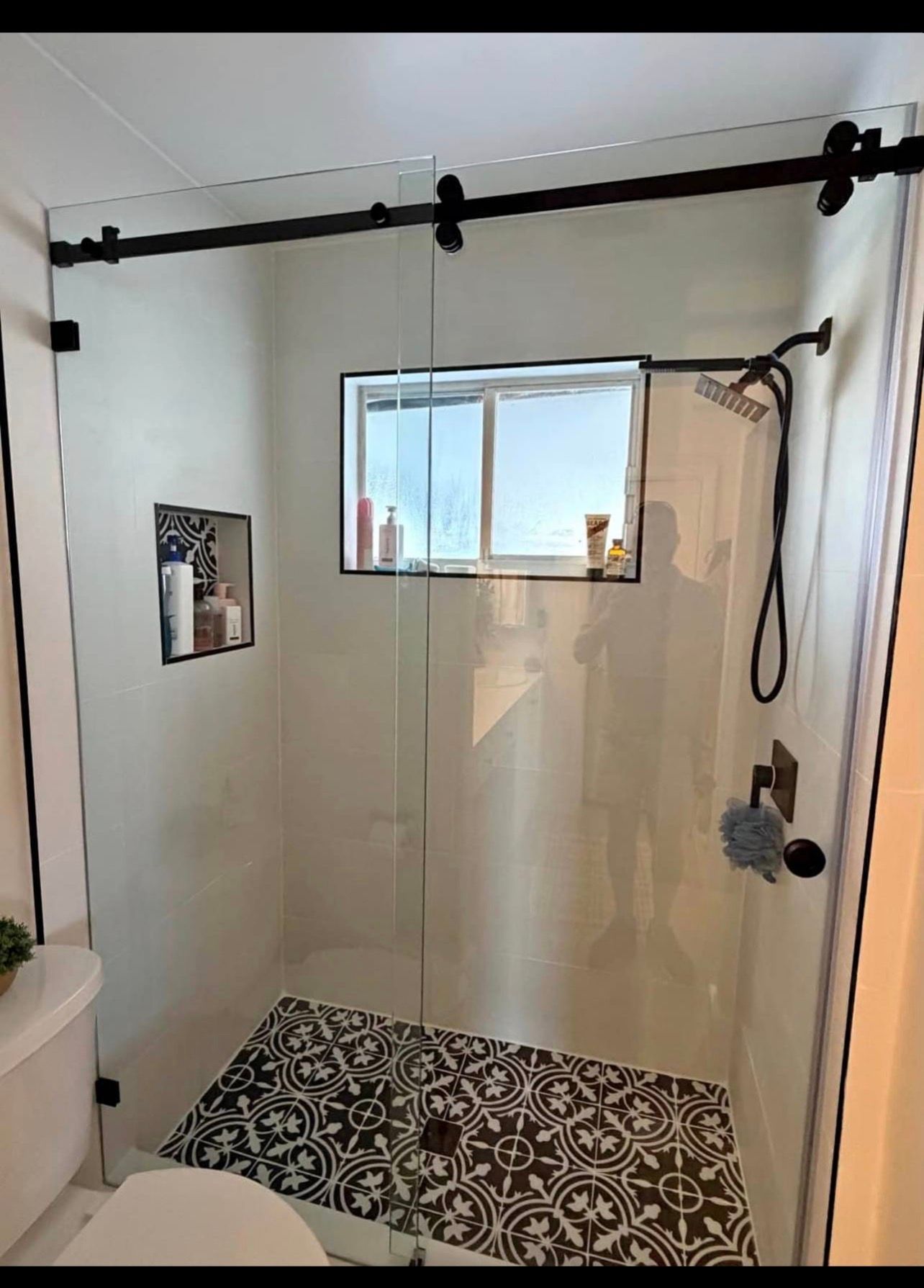 Bathroom shower with black curtain rod, frosted glass door, window, and patterned tile floor.
