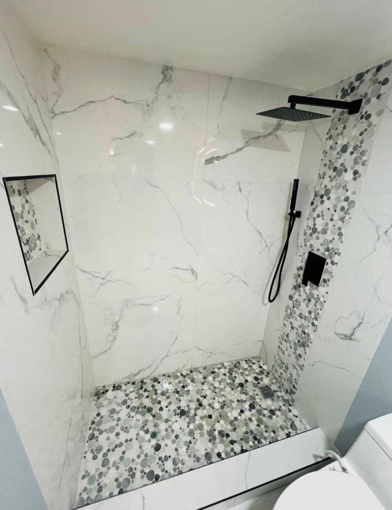 Modern shower with white marble-look tile, black fixtures, patterned floor, and wall mirror