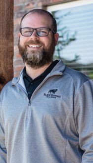 Smiling person with glasses and beard wearing a gray zip-up jacket outdoors