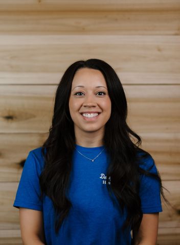 Woman with long dark hair, smiling, wearing a blue shirt, standing in front of wood paneling.