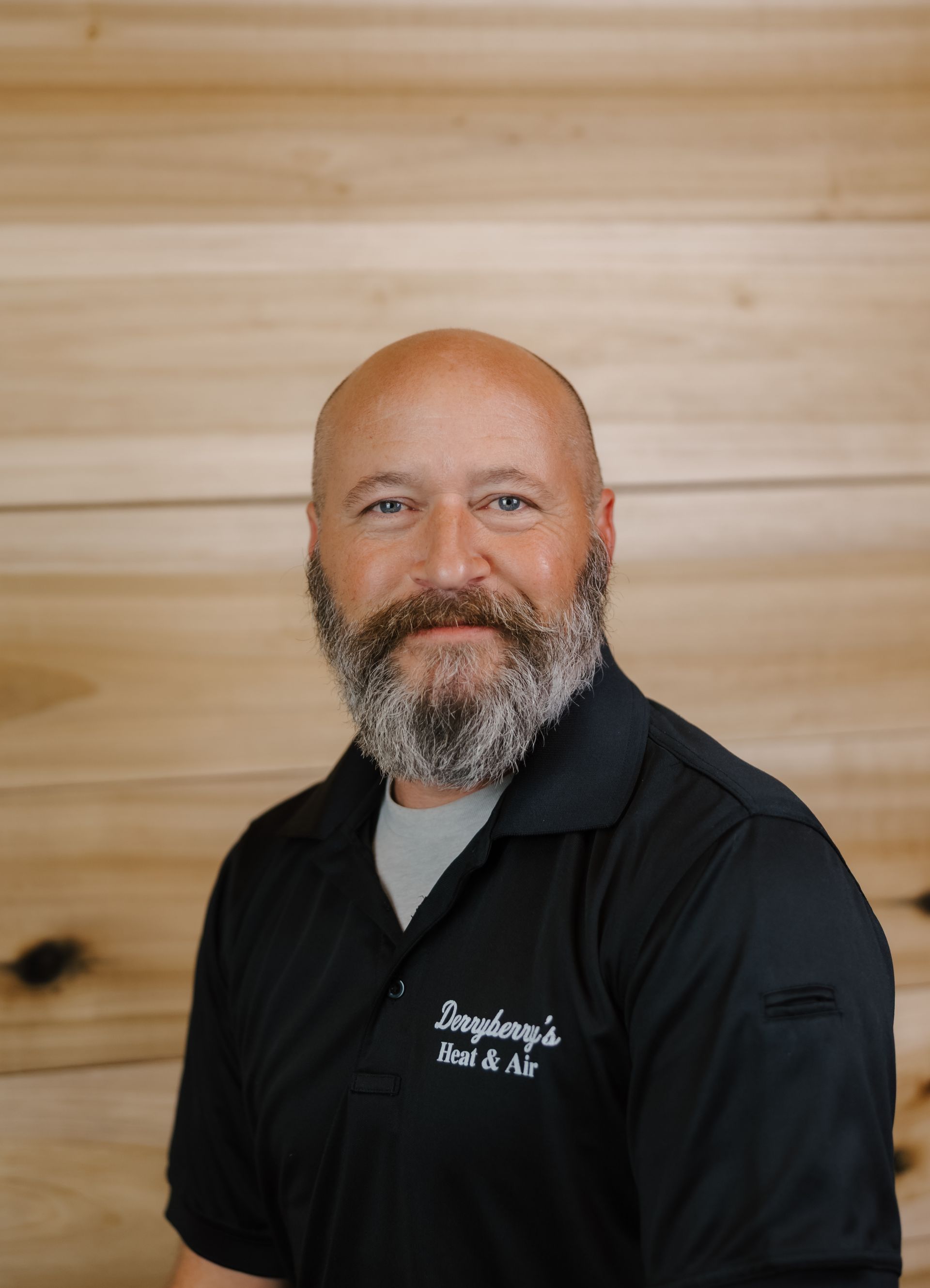 Man with a beard in a black polo shirt, standing in front of wood paneling.