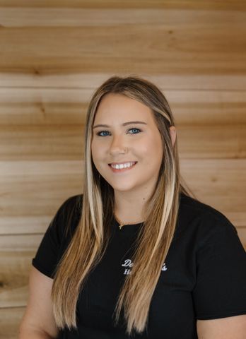 Woman with blonde hair smiling, wearing black shirt against a wood-paneled background.
