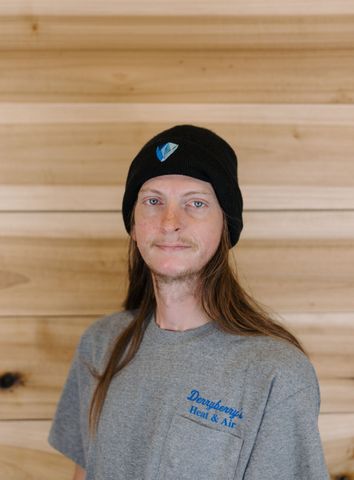 Man wearing a gray beanie and hooded sweatshirt against a wood-paneled wall, looking at the camera.