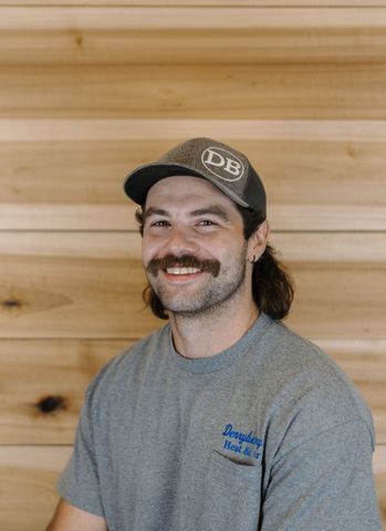 Man with glasses and a gray beanie smiles, in front of a wooden wall.