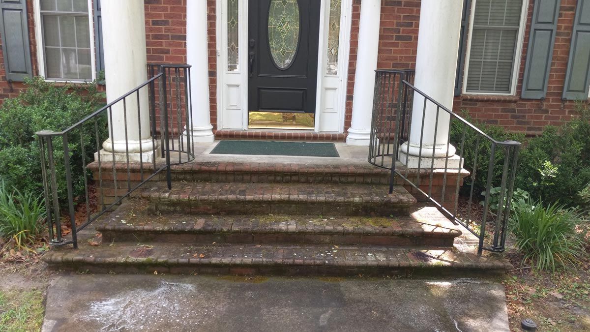 The front porch of a brick house with steps and a wrought iron railing.
