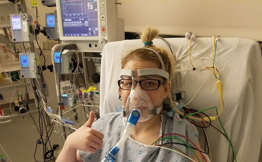 A woman wearing an oxygen mask is giving a thumbs up in a hospital bed.
