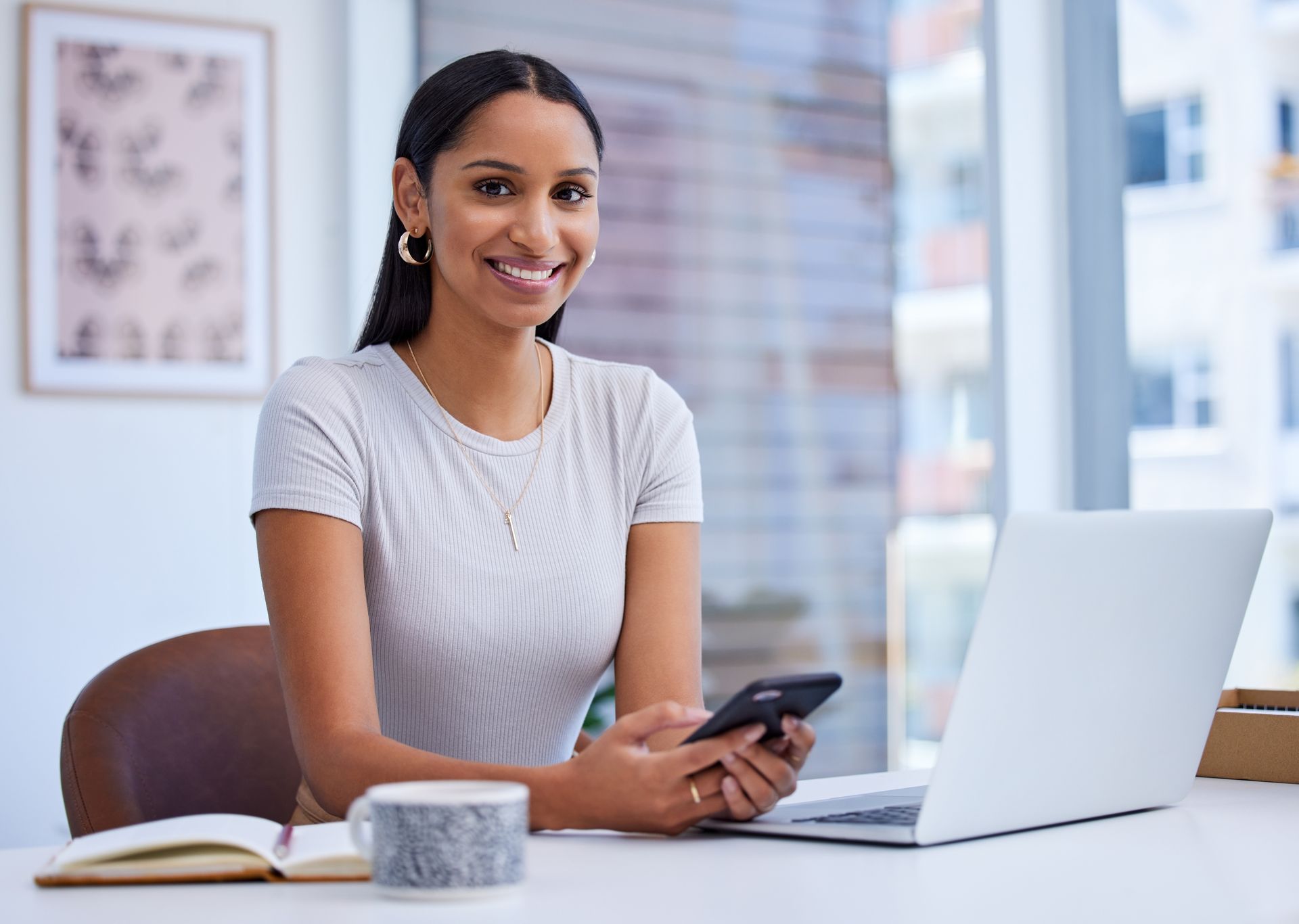 A woman is sitting at a desk with a laptop and a cell phone.