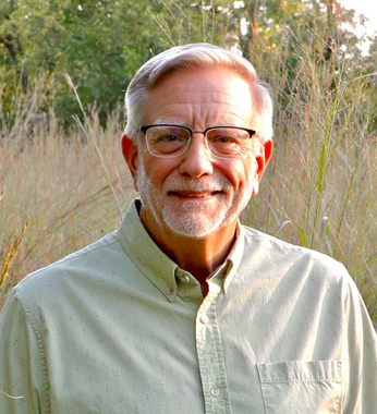 Man with gray hair and beard wearing glasses smiles in a field.