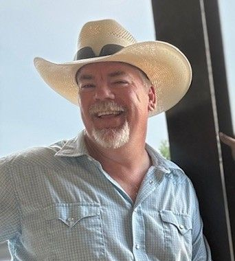 Man in cowboy hat smiles outdoors, wearing a light blue button-up shirt.