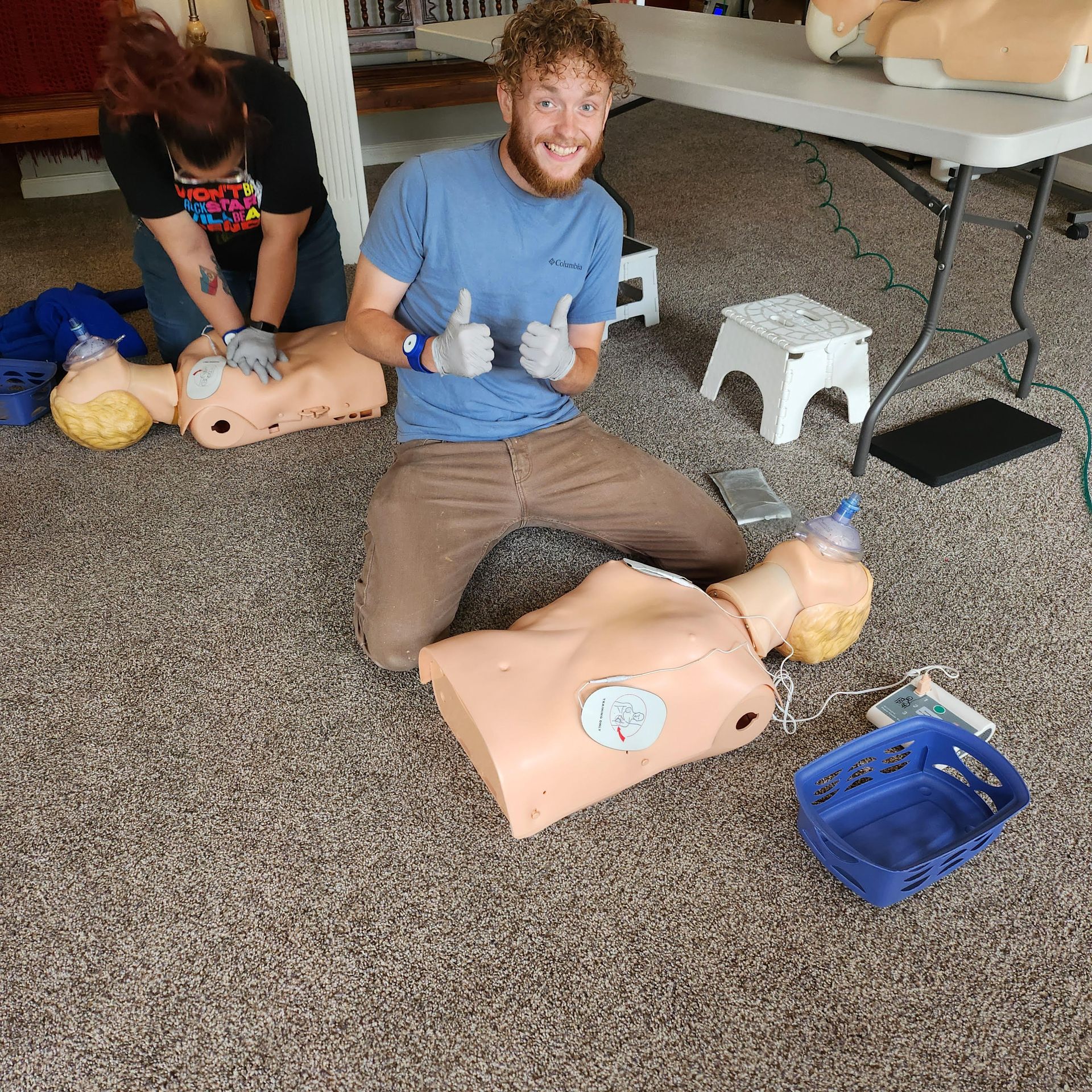 Two people practice CPR on mannequins on a carpeted floor, one giving thumbs up.