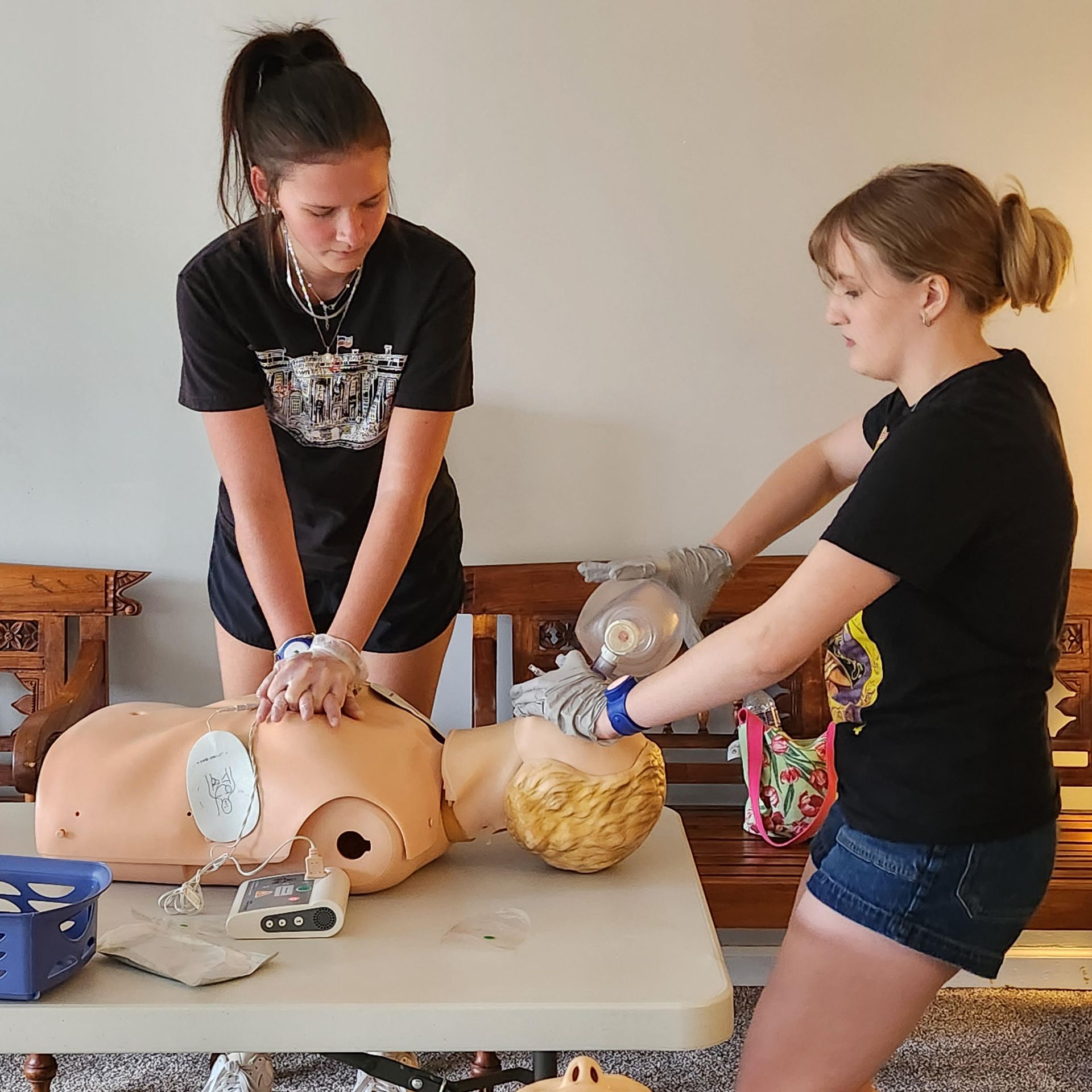 Two people practice CPR on a training manikin at a table, using a bag mask and chest compressions.