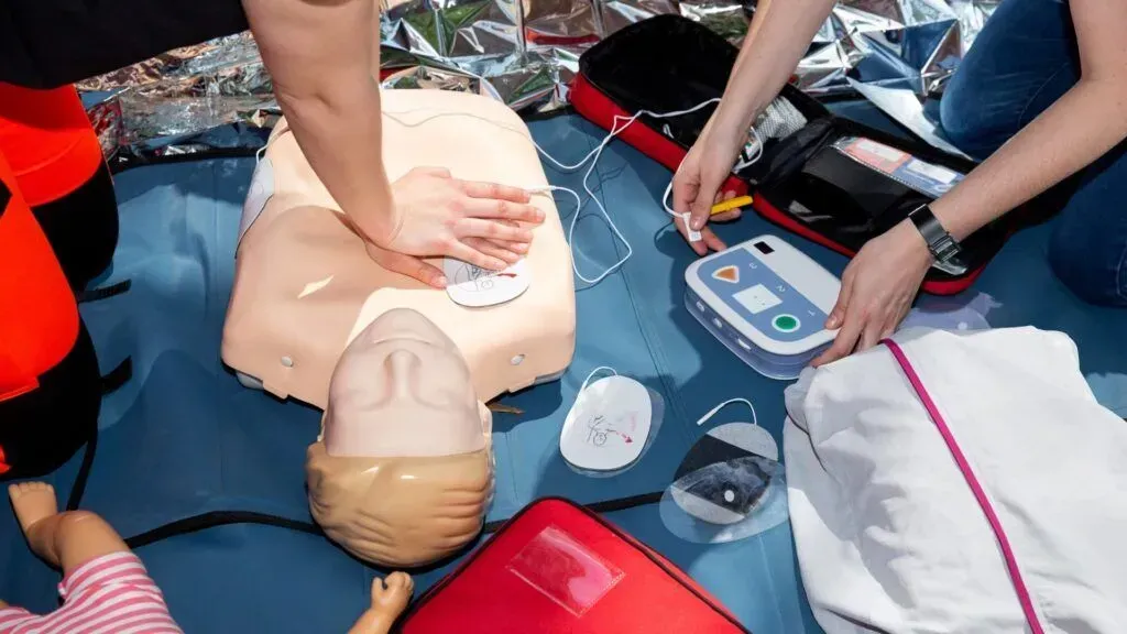 CPR training scene with a mannequin on a mat while trainees use a defibrillator and prepare supplies