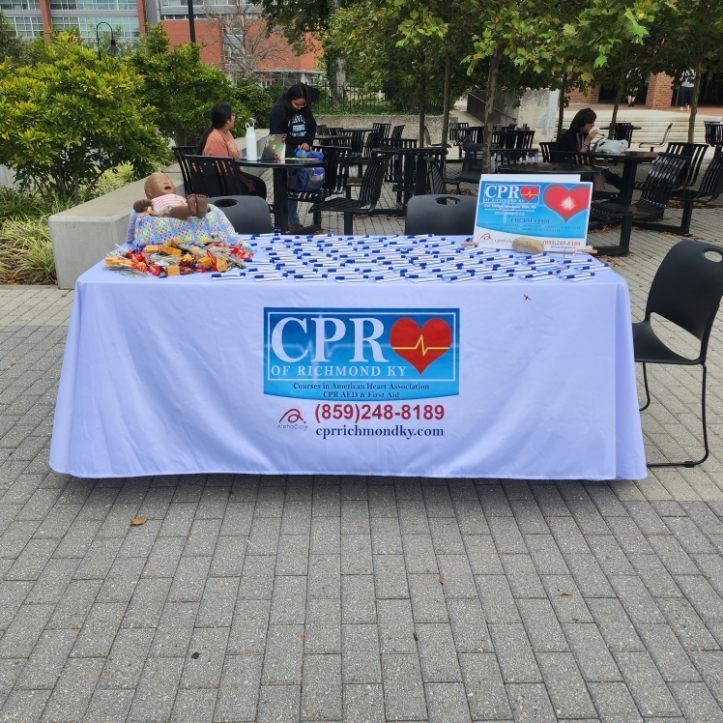 CPR table with blue tablecloth and sign in an outdoor plaza, with CPR materials displayed.