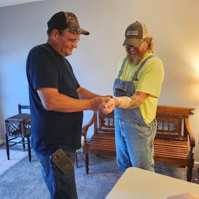 Two men hold hands in a living room, one wearing overalls and a cap.