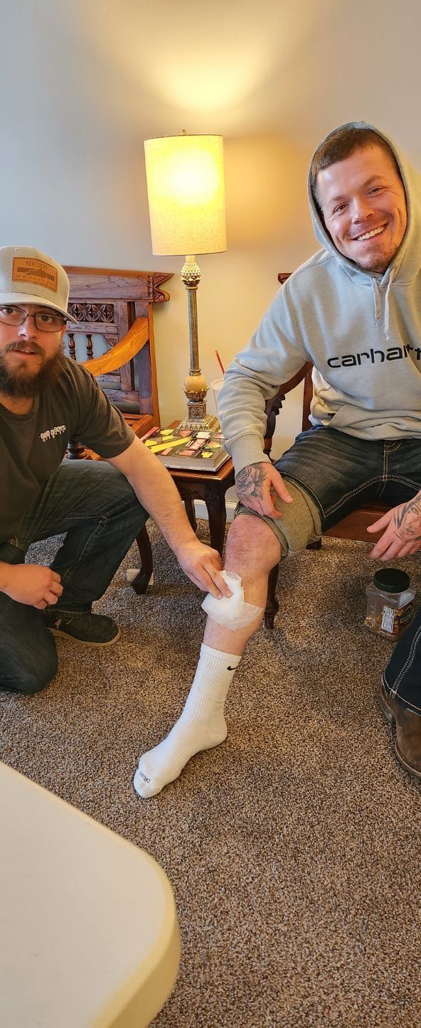 Two men sit indoors by a lamp; one smiles while wearing a white foot cast.