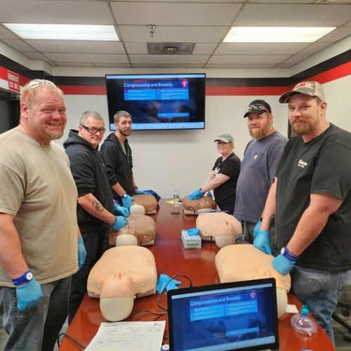 People practicing CPR on training mannequins in a classroom, with monitors and laptops on a long table.