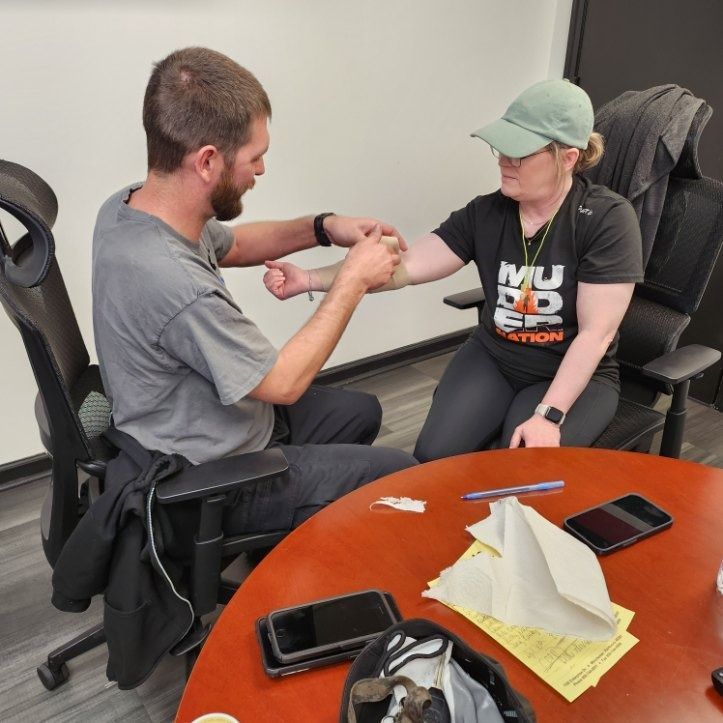 Two people in an office setting examining a wrist while seated at a table with papers and phones.