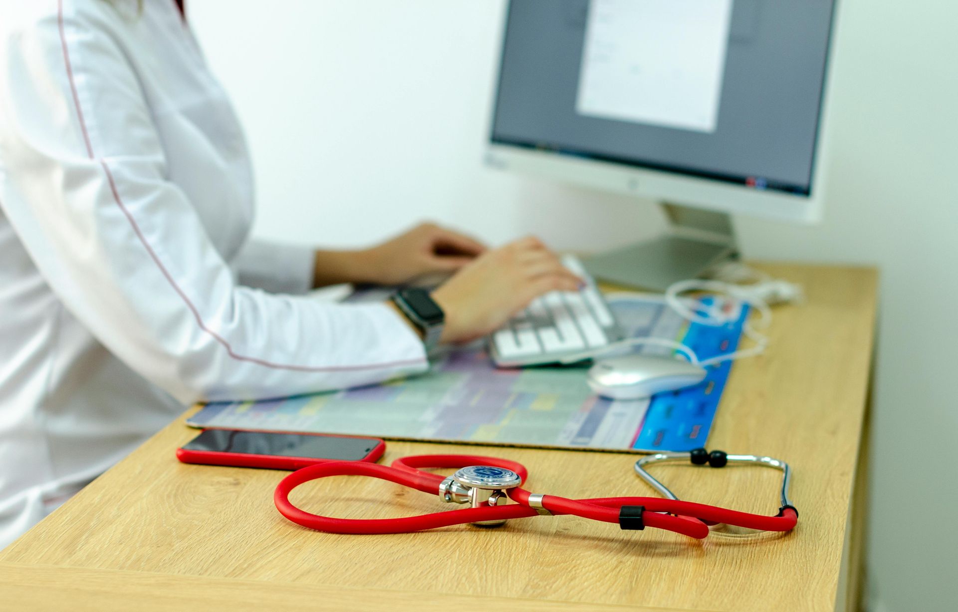 Doctor typing at a desk with a red stethoscope in front of a computer monitor