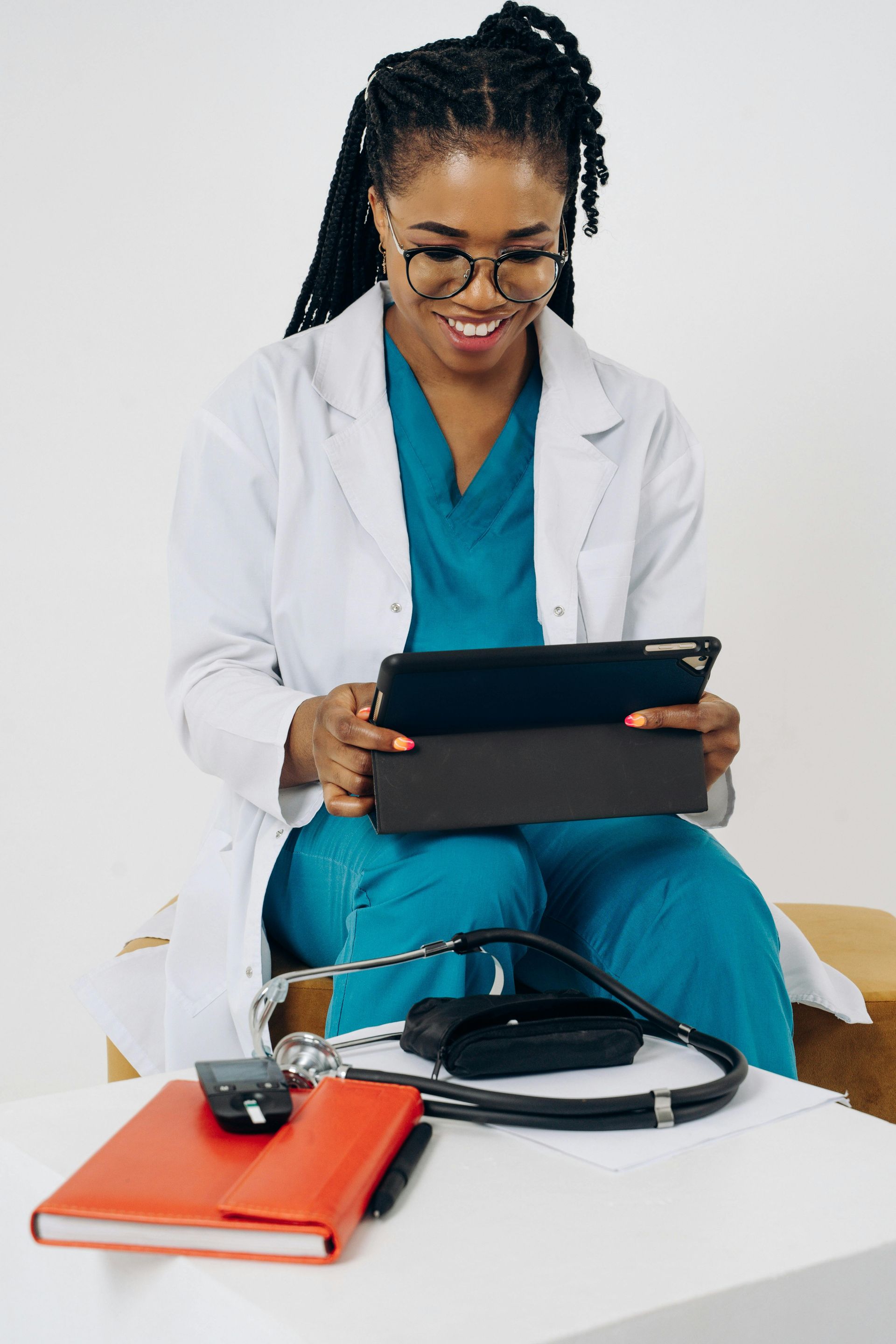 Doctor in a white coat using a tablet beside a stethoscope and red notebook on a desk