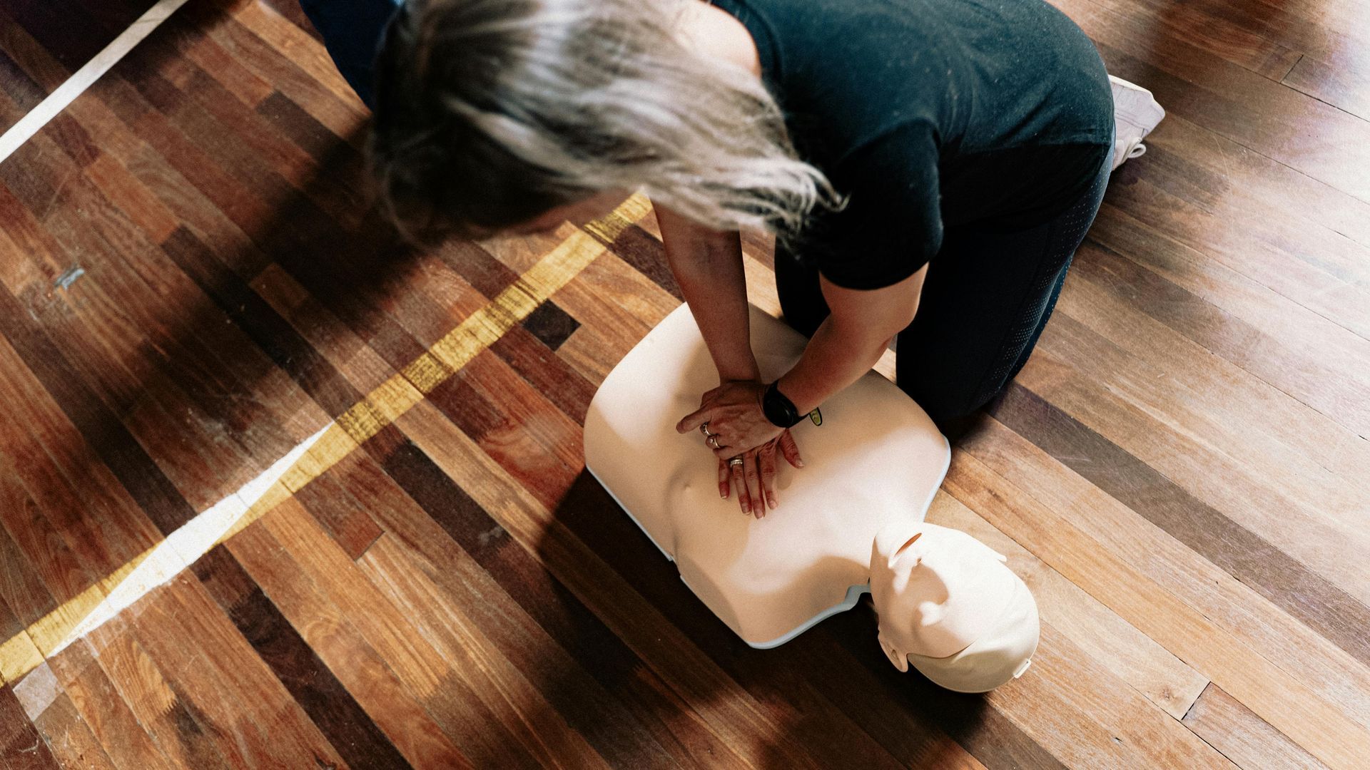 Person kneels on a hardwood floor, pressing on a CPR training mannequin's chest during practice.