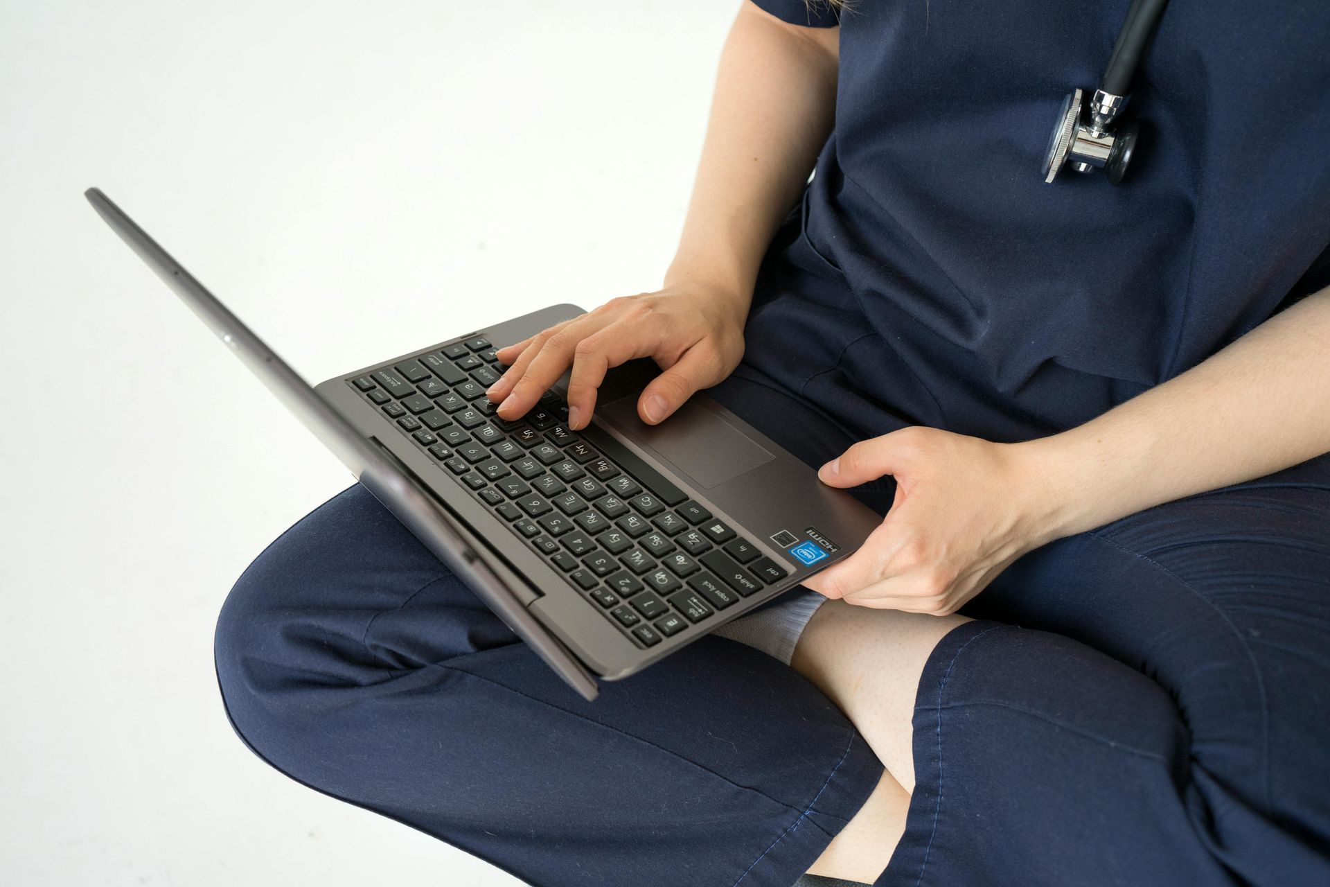 Person typing on a laptop while seated, wearing blue scrubs.
