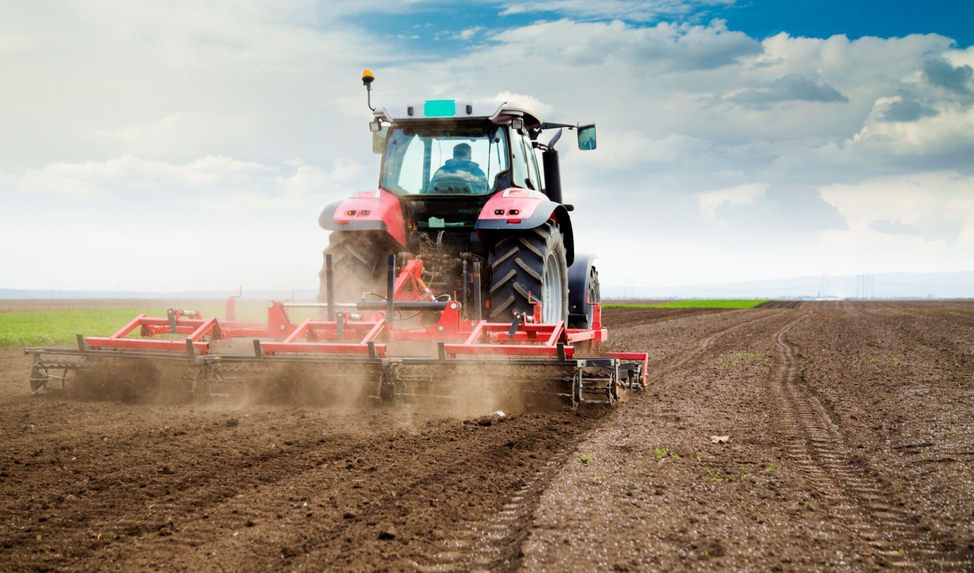 Un trattore rosso traina un attrezzo agricolo attraverso un campo arato e polveroso, sotto un cielo luminoso e nuvoloso.